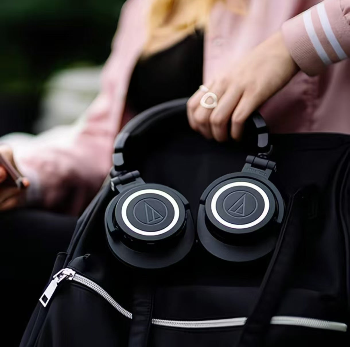 A woman holding Audio-Technica ATH-M50xENSO over-ear headphones resting on a black backpack — ideal for music lovers and professionals who need high-quality sound while commuting or traveling.