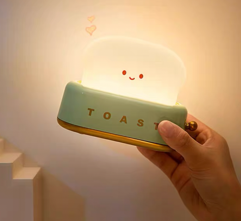Hand holding green toaster night light by the brass lever showing portable size and warm glowing toast lamp face
