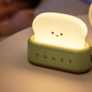 Green toaster night light glowing on bedside table beside sleeping person for soft sleep-friendly bedroom lighting