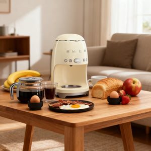The elegant Smeg 50's Retro Style glass coffee carafe sitting on a modern white kitchen countertop next to a handful of coffee beans, with a stainless steel sink and faucet in the background.