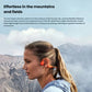 Woman hiking in mountains wearing Shokz OpenRun Pro 2 headphones, demonstrating secure fit with flexible titanium alloy hooks and open-ear design